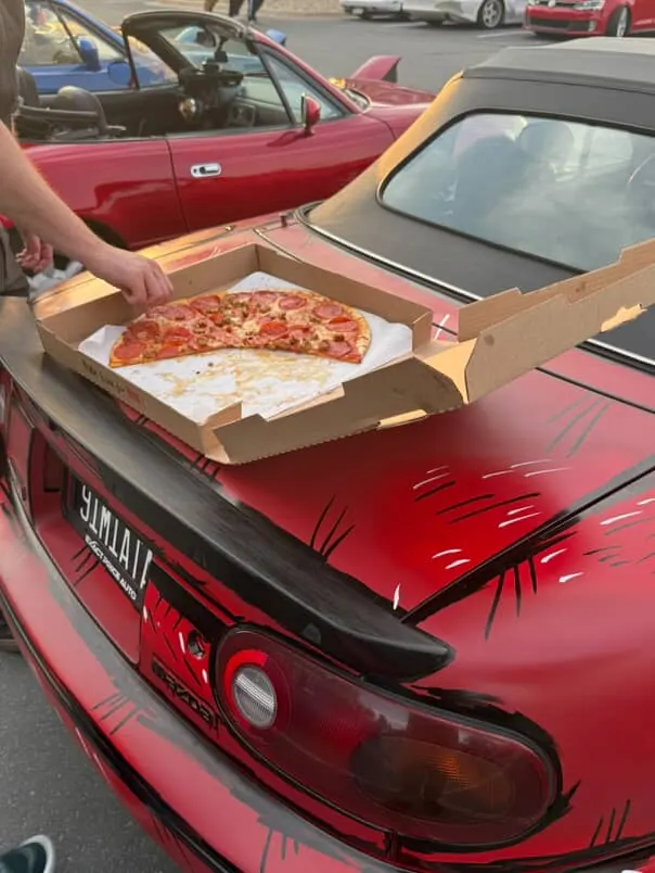 A pizza box open on the trunk of a red Miata